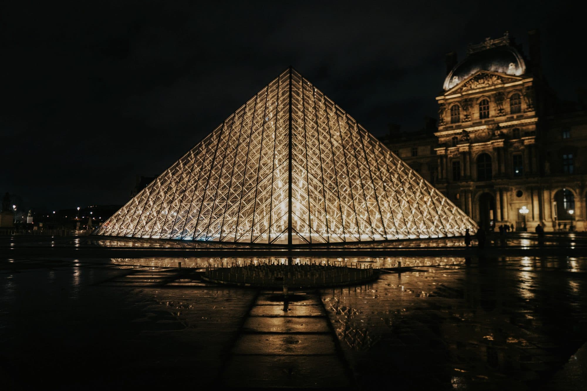 The Louvre pyramid lit at night, reflected in wet paving
